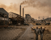 Two men walking with cattle beside closed factory buildings and a power plant with smoking chimneys